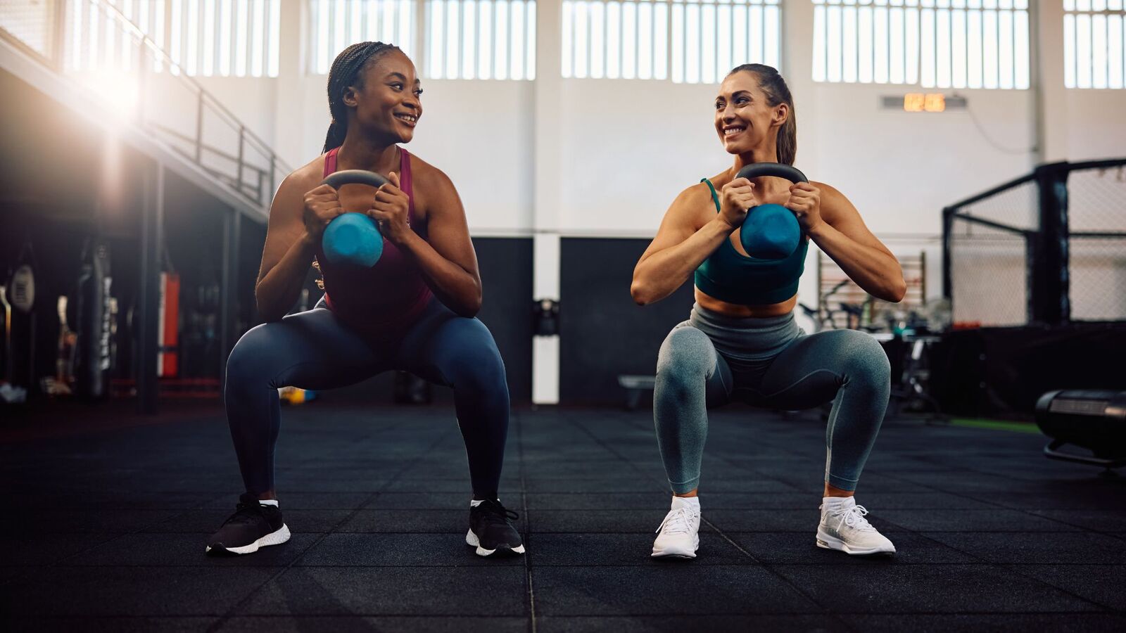 Two women laughing mid-squat with kettlebells in a bright gym